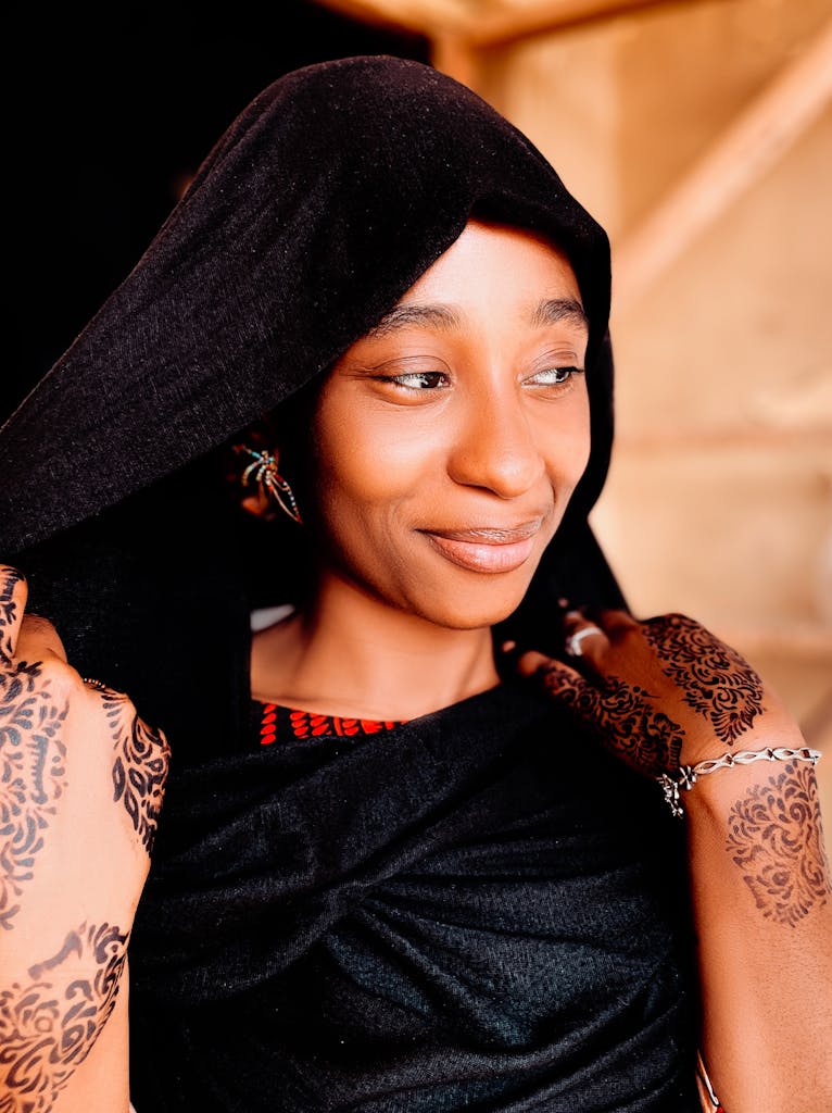 A joyful woman wearing a black shawl adorned with intricate henna designs, smiling softly.